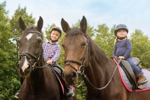 Young kids, brother and sister, 7 year young boy and 3 year young girl sitting on the back of their horses. Real People, Young kids outdoor candid horseback riding portrait.