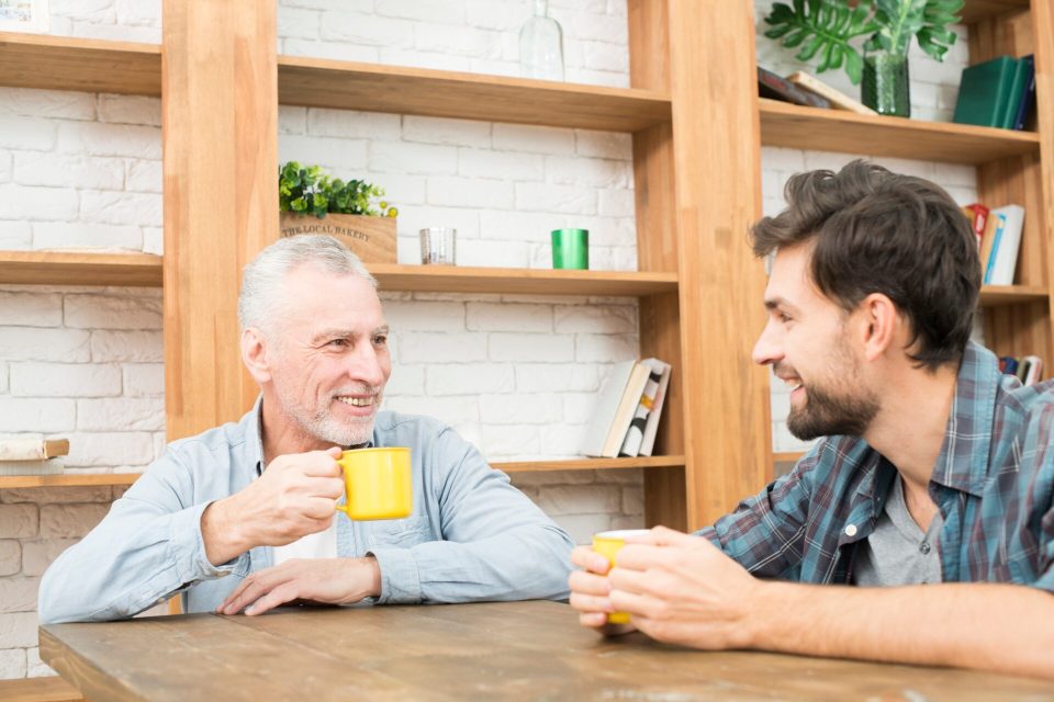 Äldre man och yngre man samtalar med kaffekoppar vid ett köksbord